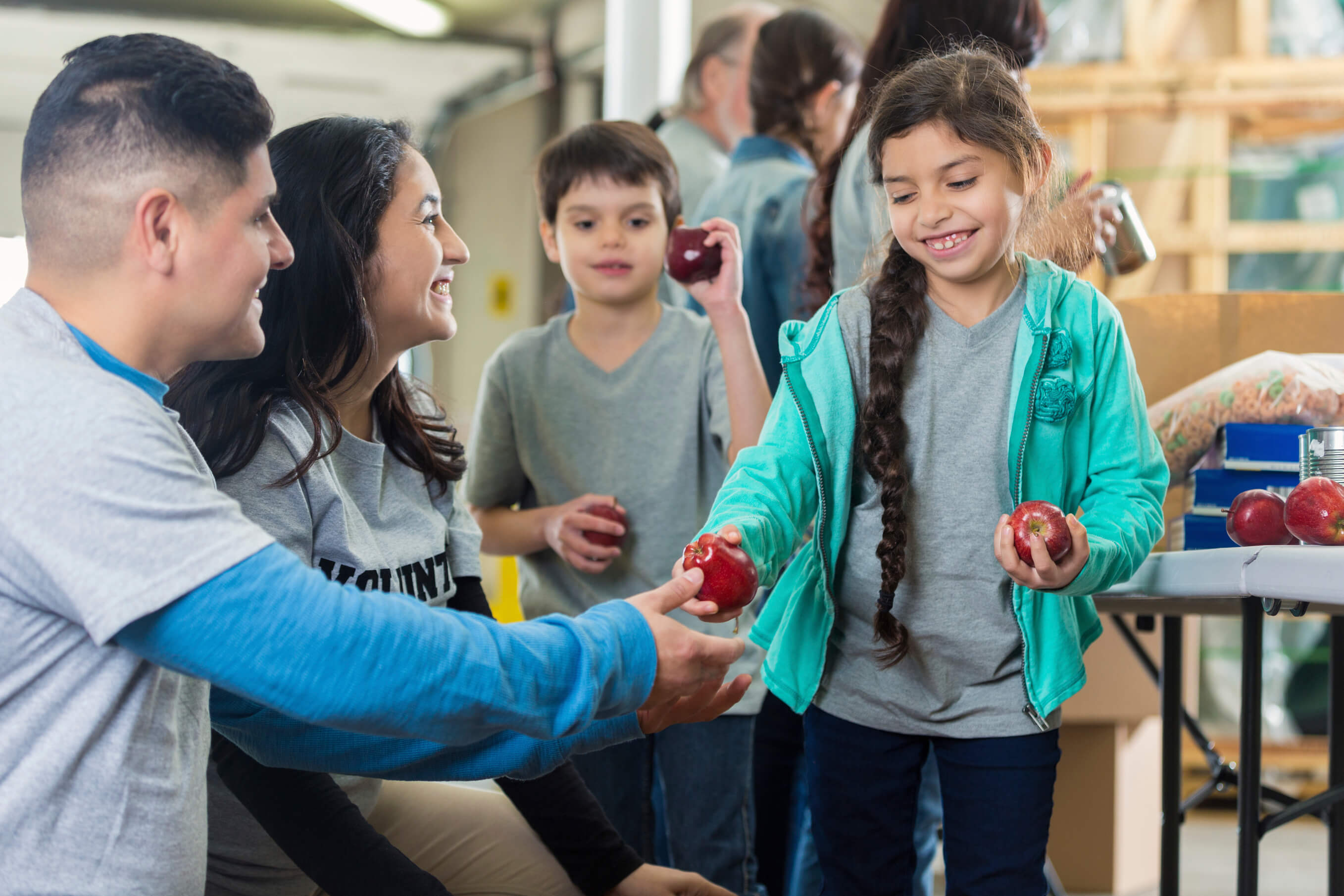 Mid adult Hispanic father and mother give their elementary age son and daughter apples in community food bank or soup kitchen. The children smile as they receive the apples. Volunteers are working in the background.